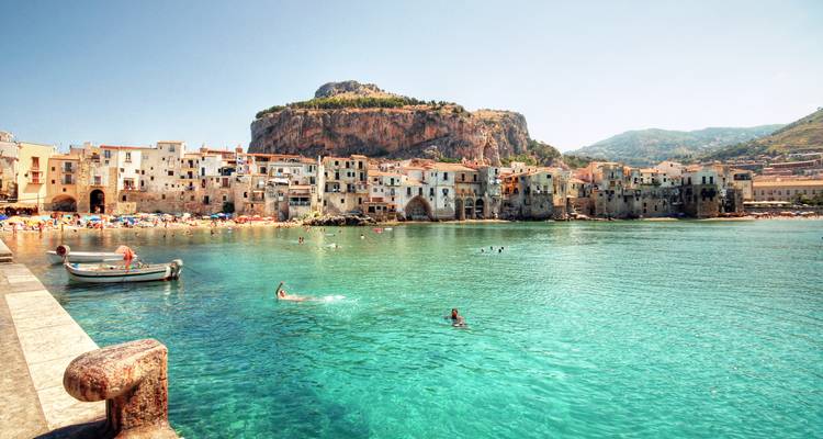 Türkisfarbene Bucht und mittelalterliche Uferpromenade von Cefalù mit Schwimmern und einem kleinen Boot unter der Sommersonne.