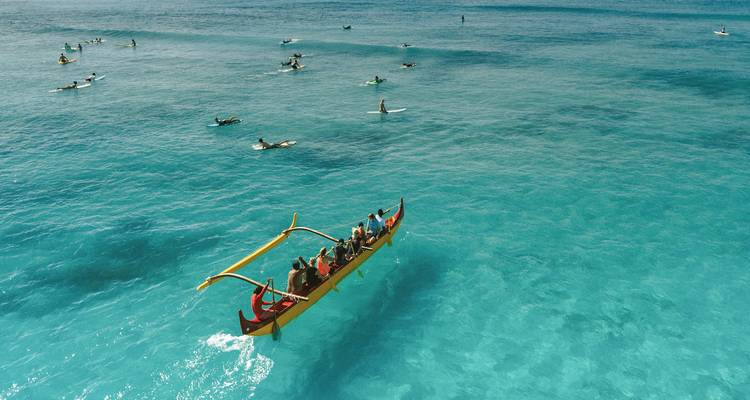 Pirogue traditionnelle à balancier avec rameurs se déplace dans une mer turquoise au milieu de surfeurs dispersés.