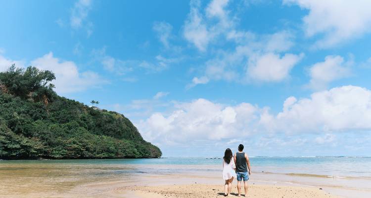 Un couple marche main dans la main le long d'une plage de sable déserte vers un promontoire couvert de jungle sous un ciel bleu.