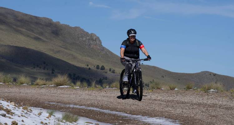 Ein Mountainbiker fährt auf einer Schotterstraße neben grasbewachsenen Hängen und Flecken von liegengebliebenem Schnee hinauf.