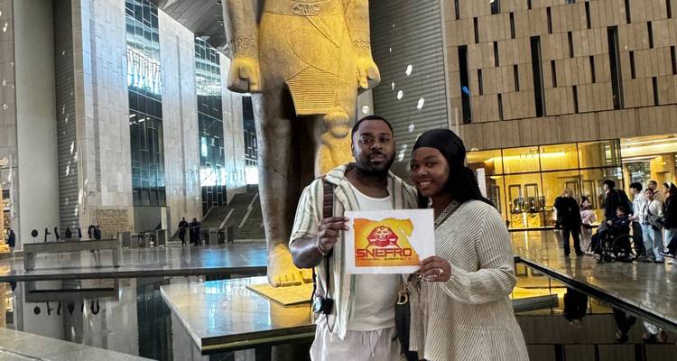 Twee mensen poseren met een bord voor een groot standbeeld.