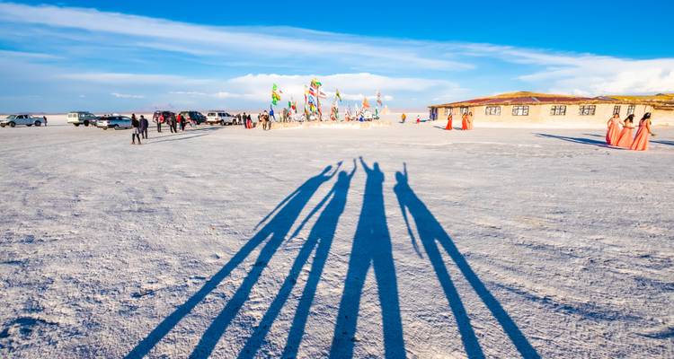 Eine Salzebene mit bunten Flaggen und langen Schatten von Menschen, Fahrzeuge sind im Hintergrund geparkt.