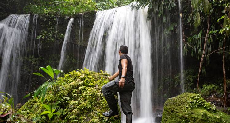 Homme debout à la base d'une cascade dans un environnement luxuriant.