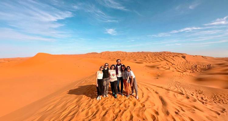 Kleine Gruppe von Reisenden, die auf hohen orangefarbenen Sanddünen unter blauem Himmel posiert.