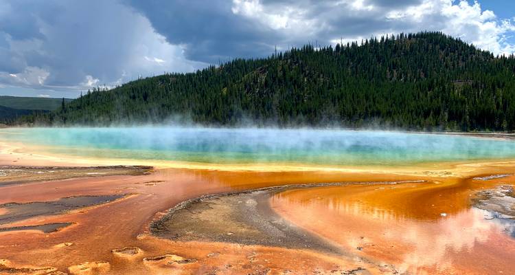 Steam rising from vibrant colored hot spring surrounded by forested hills.