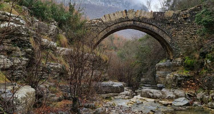Een stenen brug over een beek met kale bomen en rotsachtig terrein.