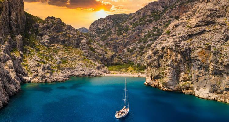Crique sereine avec un yacht à voile et paysage montagneux au coucher du soleil.