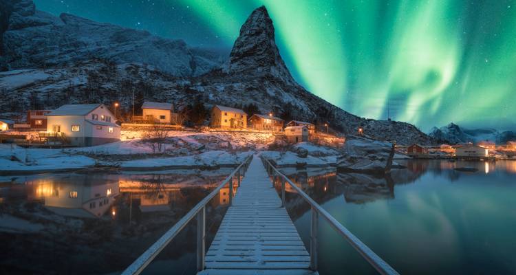 Atemberaubende grüne Nordlichter erhellen ein verschneites Fjorddorf und einen spitzen Berggipfel, die sich in ruhigem Wasser spiegeln.