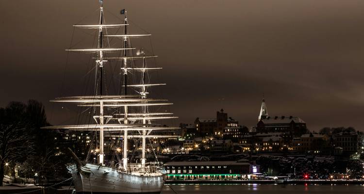 Een nachtelijk zicht op een zeilschip aangemeerd bij een stadsgezicht.