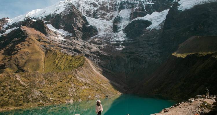 Personne debout près d'un lac glaciaire avec des montagnes enneigées.