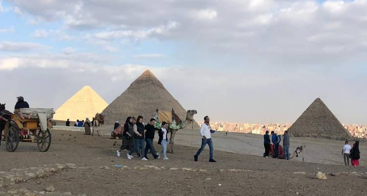 Des gens marchant près des pyramides de Gizeh, avec des chameaux et une charrette.