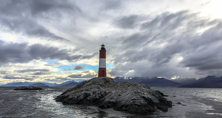 Phare rouge et blanc se dressant sur un îlot rocheux dans le canal de Beagle sous un ciel nuageux dramatique et des montagnes enneigées au loin