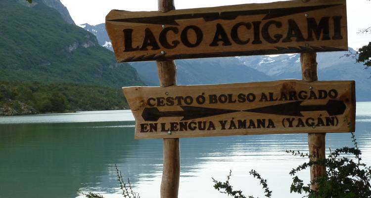 Panneau en bois indiquant 'Lago Acigami' au bord d'un lac turquoise calme entouré de forêt et de pics enneigés