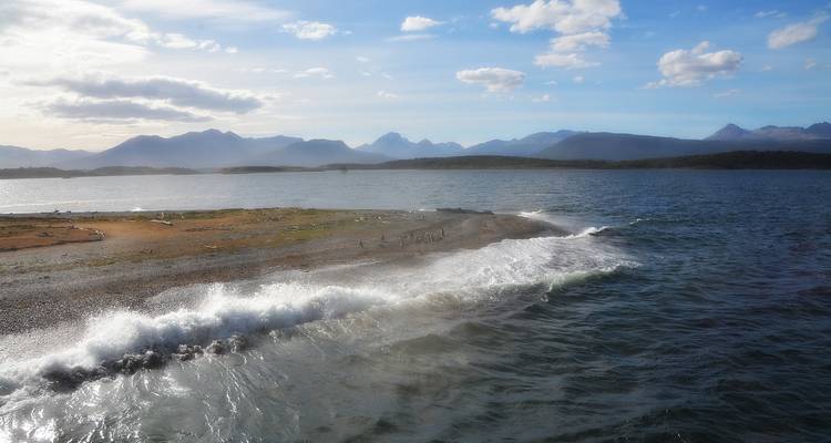 Vagues se brisant contre un banc de sable bas s'avançant dans un large chenal avec des montagnes brumeuses au loin