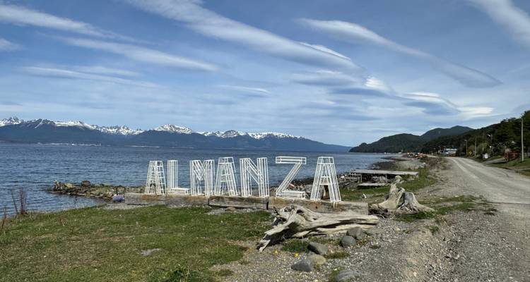 Escena costera al borde de la carretera con grandes letras de alambre que forman 'ALMANZA' contra el Canal Beagle y picos nevados.