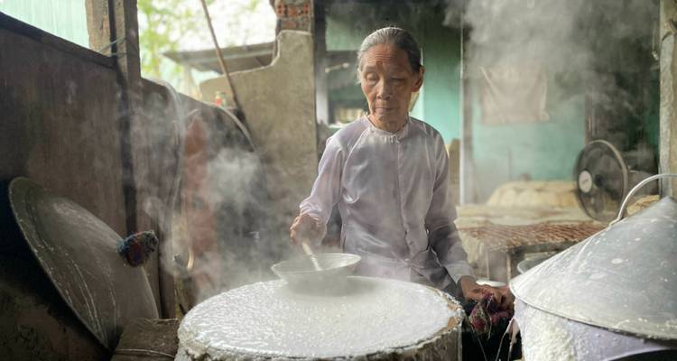 Femme âgée cuisinant de la nourriture traditionnelle dans un cadre rustique.