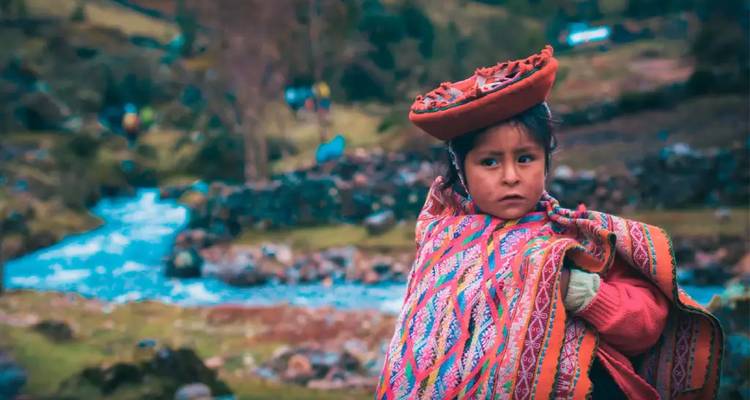 Niño con vestimenta tradicional peruana de pie junto a un arroyo y paisaje rocoso.