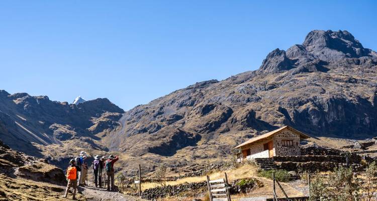 Excursionistas en un sendero de montaña con una cabaña rústica y picos rocosos.