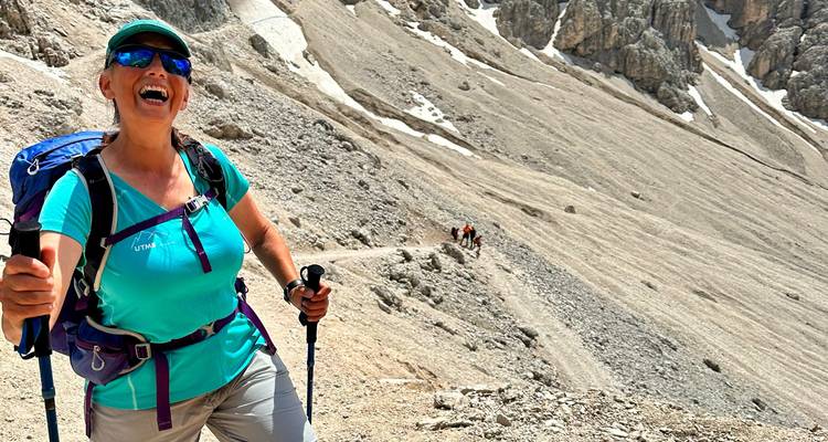 Glimlachende wandelaar met trekkingstokken loopt omhoog over een kaal rotsachtig pad met Dolomieten-pieken op de achtergrond.