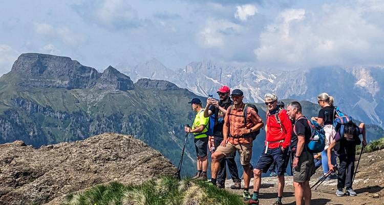 Rij wandelaars pauseren op een rotsachtig uitkijkpunt met gelaagde Dolomieten-bergkammen erachter.