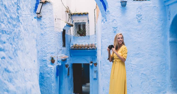 Frau in hellgelbem Kleid lächelt, während sie eine Kamera in Chefchaouens blauer Straße hält.