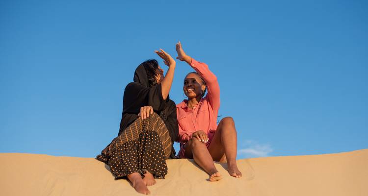 Zwei Personen sitzen auf einer Sanddüne unter einem klaren blauen Himmel.