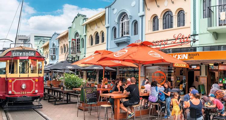 Scène de rue avec tramway, bâtiments colorés, et personnes qui dînent dehors.