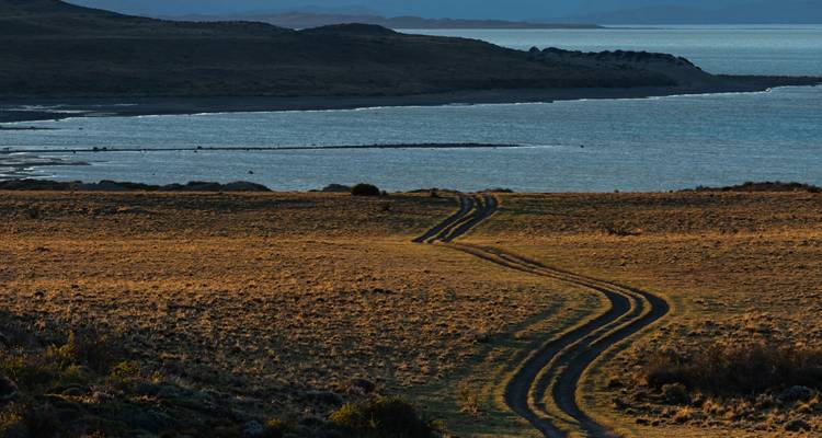 Piste de terre sinueuse traversant une steppe aride menant vers un littoral isolé et une eau bleue calme dans la lumière du soir