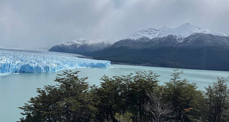 De torenhoge blauwe ijsmassa van de Perito Moreno-gletsjer ontmoet een rustig meer met sneeuwbedekte bergtoppen erachter.