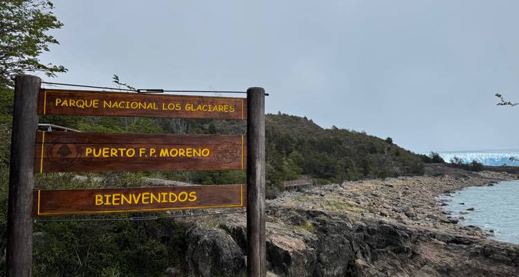 Houten welkomstbord voor Parque Nacional Los Glaciares met rotsachtige oevers en gletsjer in de verte.