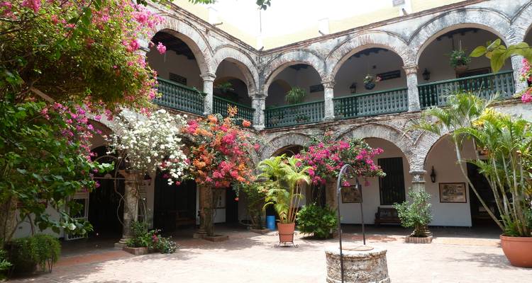 Cour coloniale luxuriante avec bougainvilliers en fleurs, arches, balcons et un puits en pierre décoratif au centre.