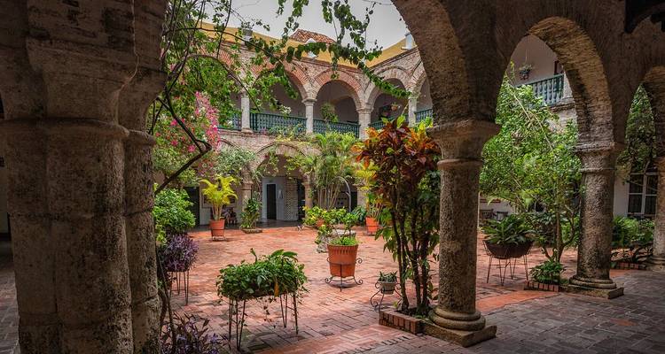 Une cour coloniale pittoresque avec un dallage en brique, des plantes en pot luxuriantes et des colonnades en pierre cintrée.