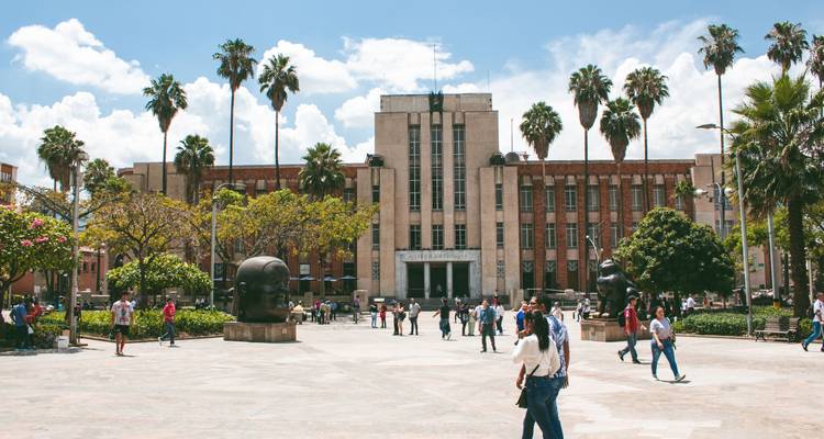 Plaza soleada con edificio del museo, palmeras y grandes esculturas de Botero, personas paseando alrededor.