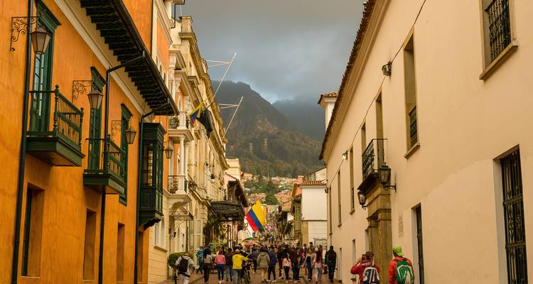 Calle colonial en el distrito histórico de Bogotá con bandera colombiana, fachadas coloridas y montañas brumosas al fondo.