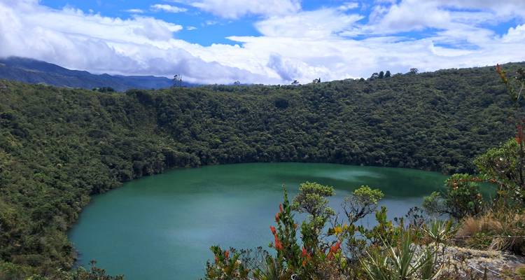 Lac de cratère vert émeraude entouré de pentes forestières densément boisées sous un ciel partiellement nuageux.