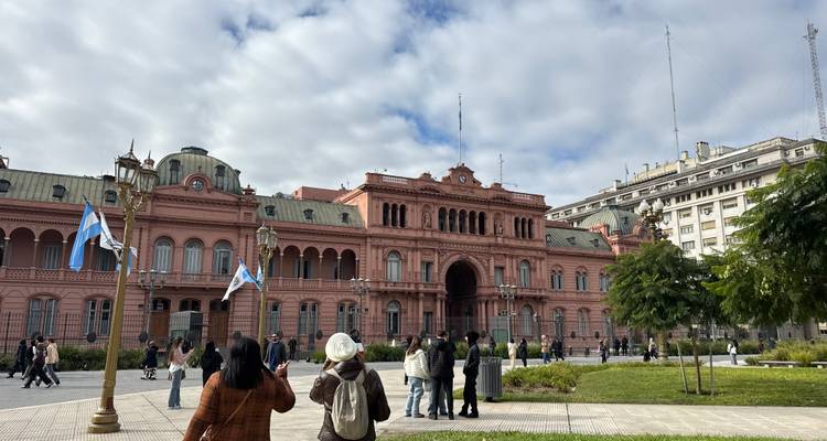 Breder pleinscène met toeristen die Casa Rosada fotograferen vanaf het zijgrasveld onder een helderblauwe lucht met wolken.