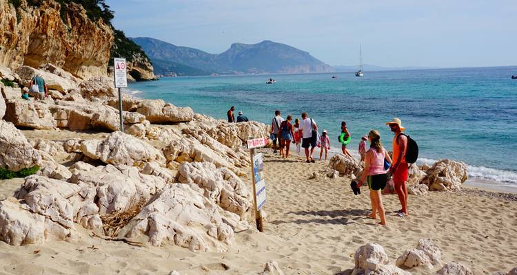 Bezoekers wandelen langs een zandstrand met hier en daar rotsformaties, op de achtergrond bergen en boten.