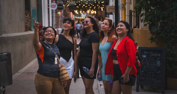 Un groupe de jeunes femmes dirigées par un guide regardent avec admiration vers le haut sous des guirlandes lumineuses scintillantes dans une rue coloniale.