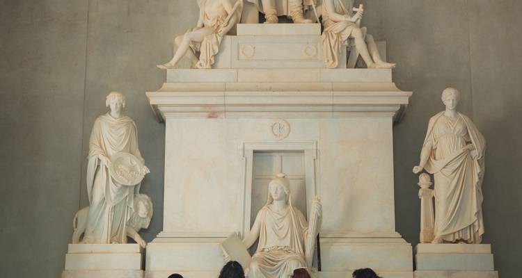 Visitors admire an ornate marble monument with classical statues inside a museum hall.