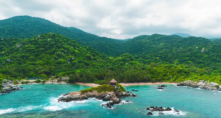 Wide aerial of lush jungle-covered headlands meeting vibrant turquoise waves in Tayrona National Park.