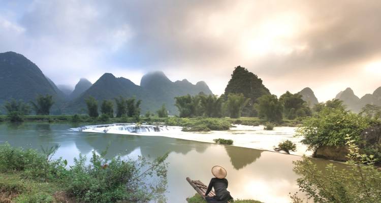 Une personne assise au bord d'une rivière avec une végétation luxuriante et des montagnes karstiques.
