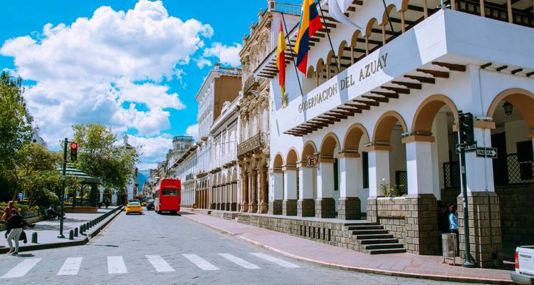 Calle histórica en Cuenca con arcos, banderas nacionales y vehículos clásicos bajo un cielo brillante
