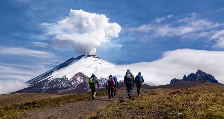 Grupo de excursionistas acercándose al volcán Cotopaxi humeante por un sendero de tierra bajo un cielo azul