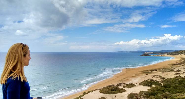 Vrouw kijkt uit over een lang, verlaten zandstrand en turquoise mediterrane wateren onder verspreide wolken.
