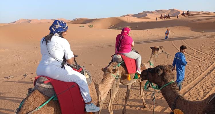 Des voyageurs assis sur des chameaux avec des couvertures colorées traversant les dunes sablonneuses marocaines sous un ciel dégagé.