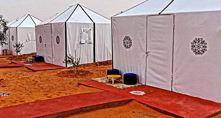 Rangées de tentes de glamping désertiques en toile blanche avec des motifs décoratifs installées sur des allées de sable recouvertes de tapis rouges.