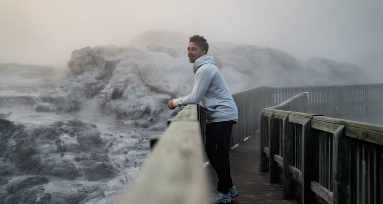 Persoon leunend op een houten reling met dampend geothermisch landschap.