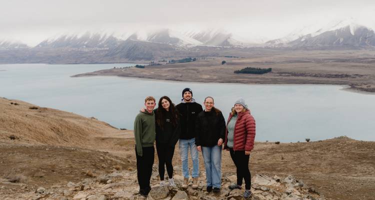 Groep van vijf mensen die op een rotsachtige heuvel staan met een watermassa en bergen op de achtergrond.