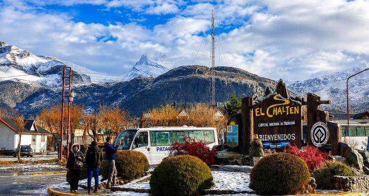 Welkomstbord van El Chaltén omlijst door sneeuwbedekte toppen en vrolijke bezoekers die naast tourbussen poseren.