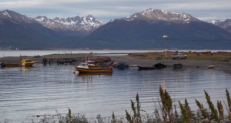 Un bateau de pêche jaune ancré dans une baie calme avec des montagnes enneigées qui s'élèvent en arrière-plan sous un ciel nuageux.